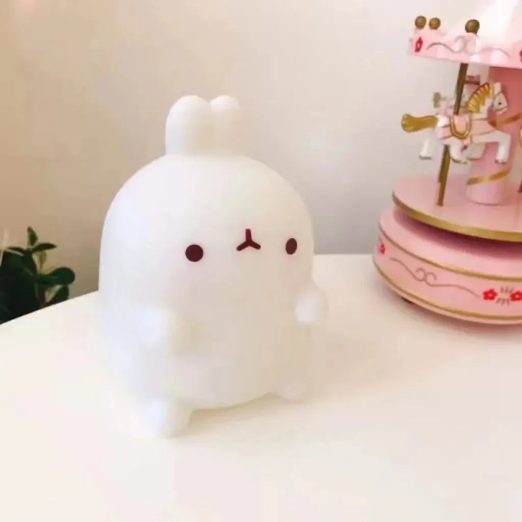 White plush toy with a simple face design on a white surface with a pink decorative item in the background.